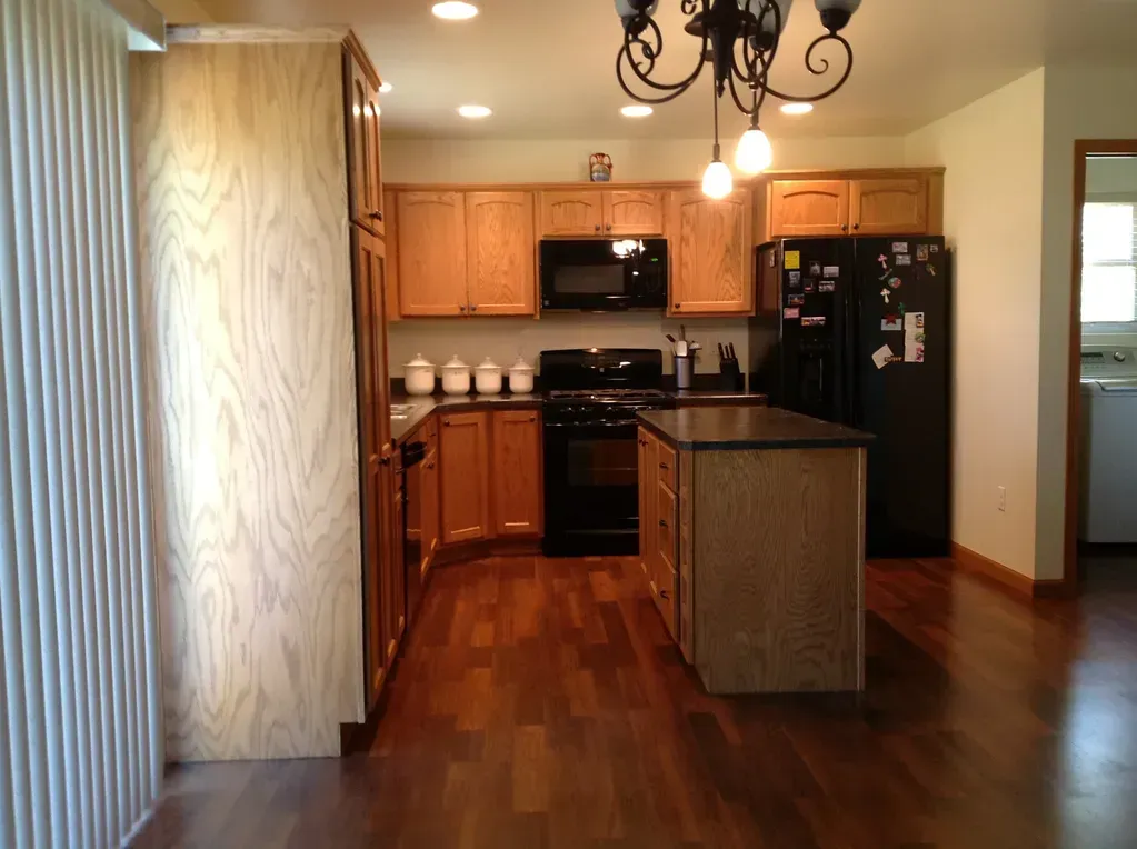 A kitchen with wood cabinets, black appliances, an island, and wood floors under a chandelier.