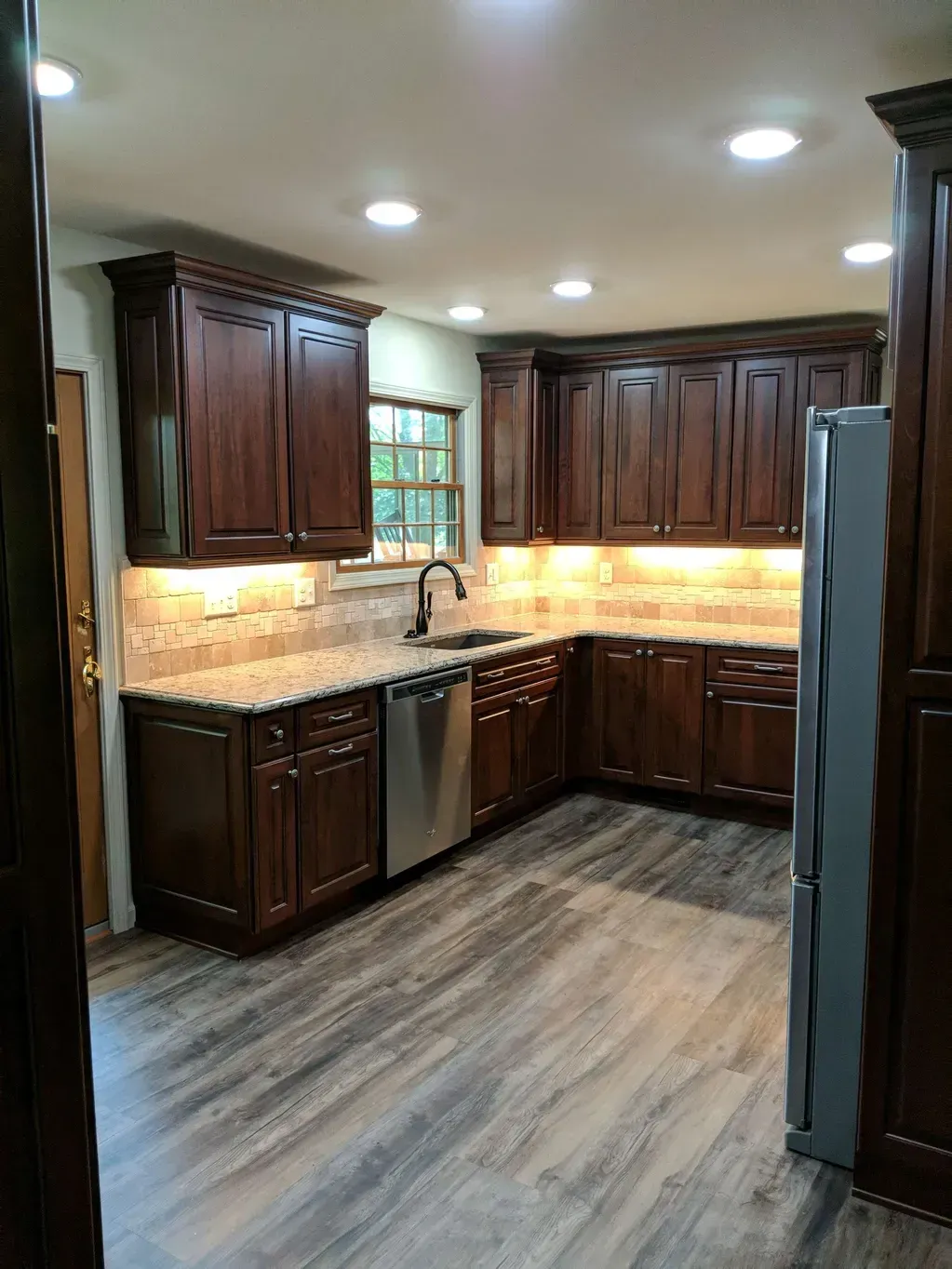 A renovated kitchen featuring dark wood cabinets, light granite countertops, stainless steel appliances, and wood flooring.