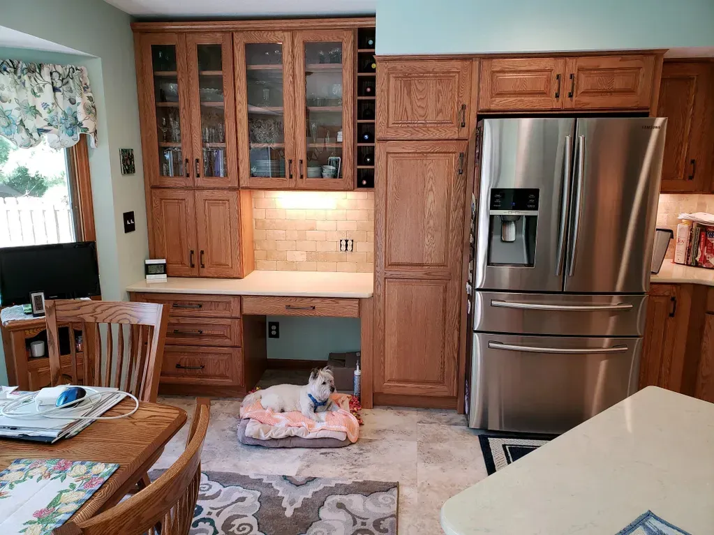 A small white dog rests on a pet bed beneath a kitchen desk flanked by wooden cabinets and a stainless steel refrigerator.