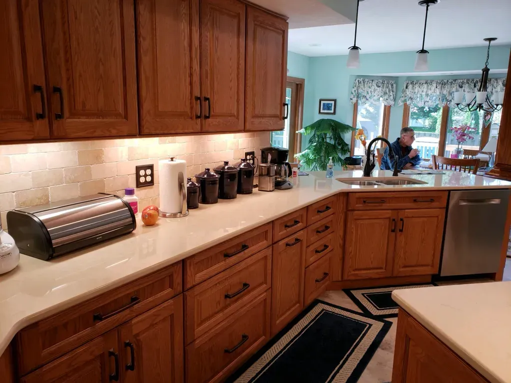 A kitchen features wood cabinets, light countertops, an L-shaped counter, and a view into a dining area with a person.