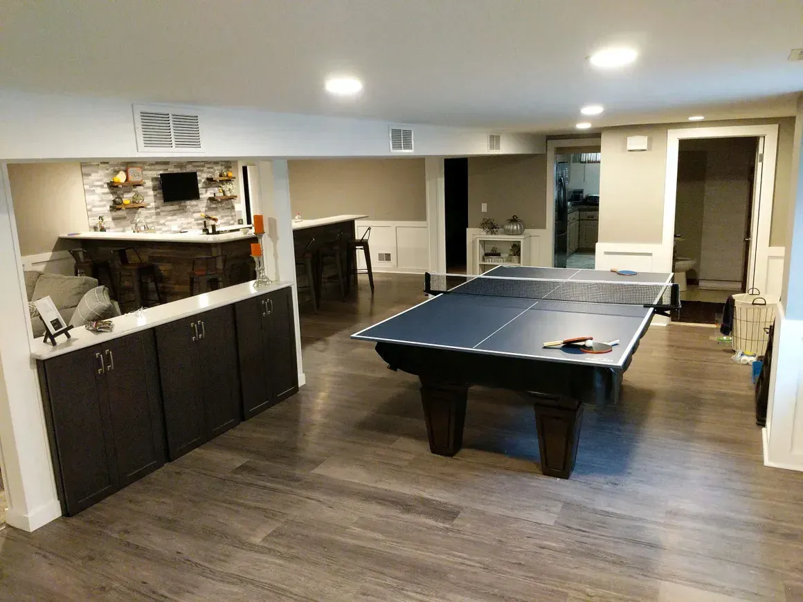 A finished basement features a ping-pong table in the center, dark wood cabinets, a bar area, and recessed lighting.