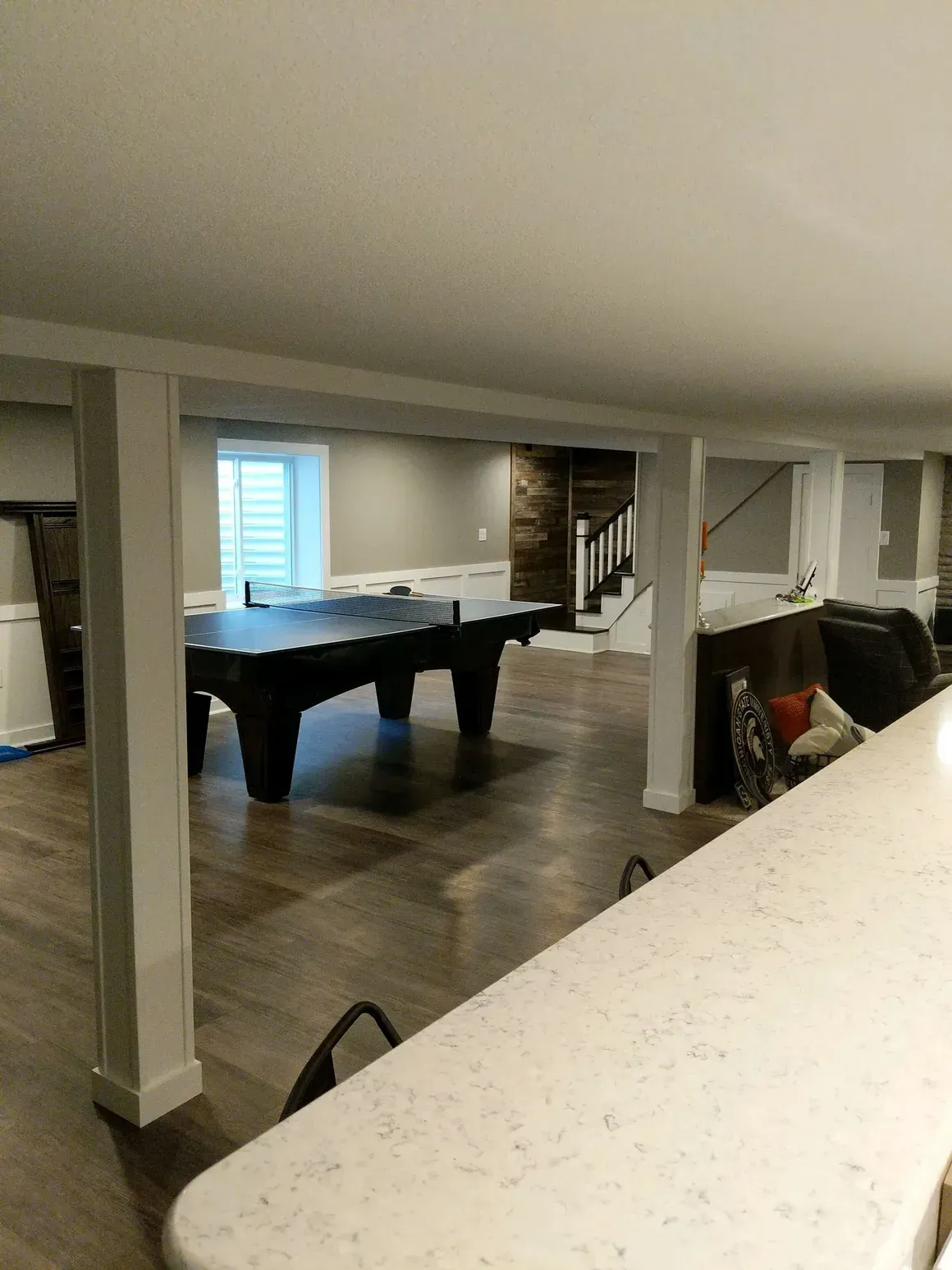 A basement recreation room featuring a ping pong table, wood floors, white support pillars, and a white countertop.