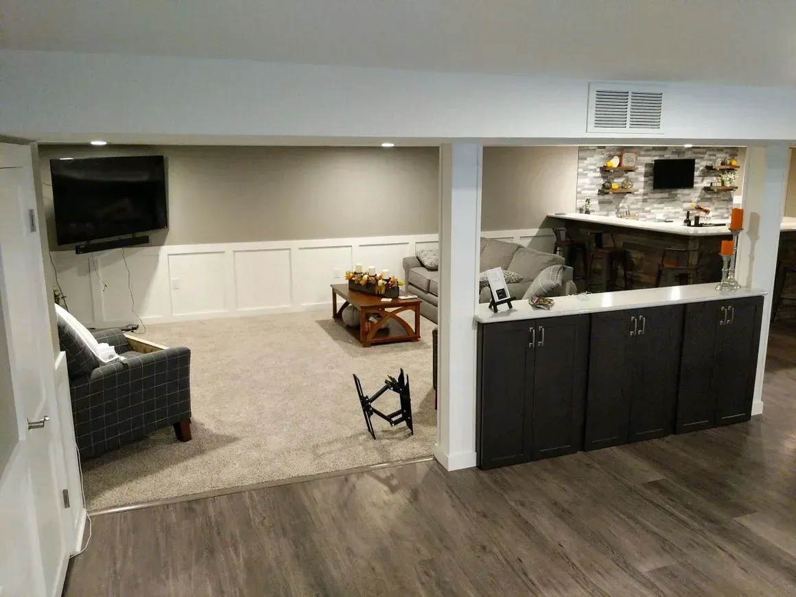 A carpeted basement with a seating area, corner television, and a dark wood bar counter with stools.