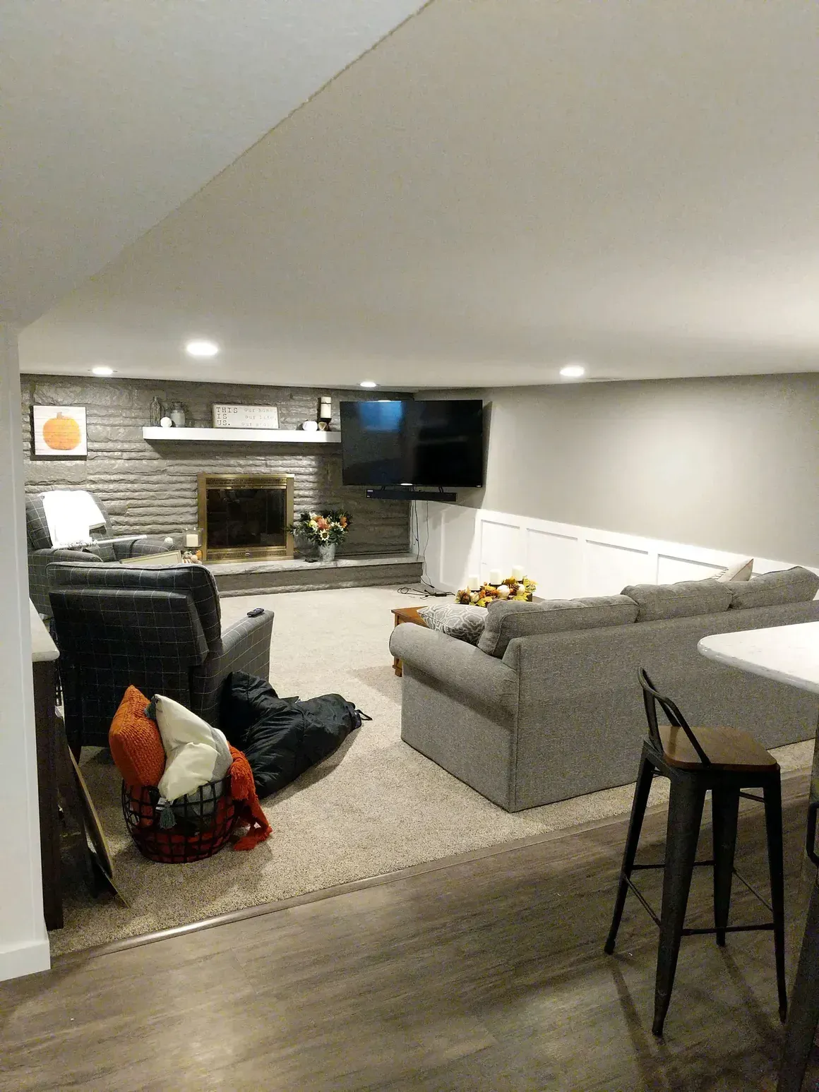 A basement living room with a brick fireplace, a gray sectional sofa, a recliner, and a wooden bar stool in the foreground.