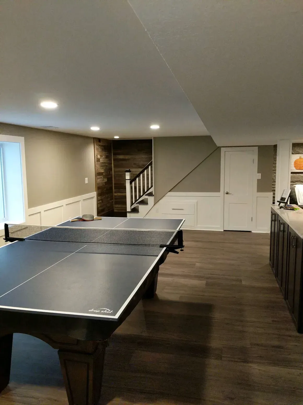 A basement interior with a ping pong table in the foreground, wood-look flooring, wainscoting, and a staircase.