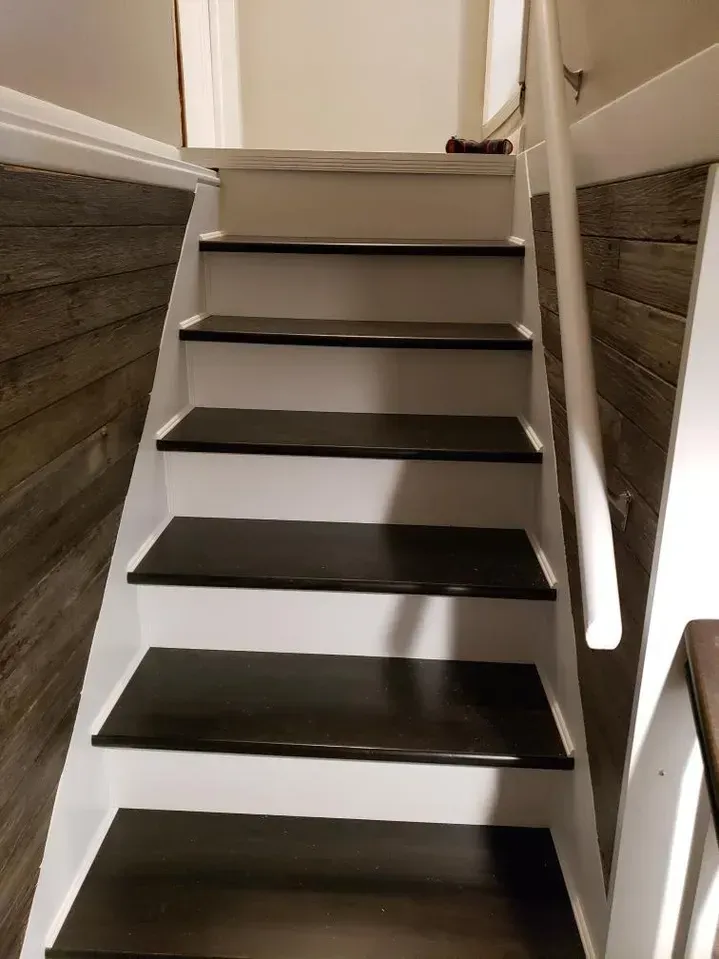 A staircase with dark brown treads and white risers, framed by rustic wood-paneled walls and a white handrail.
