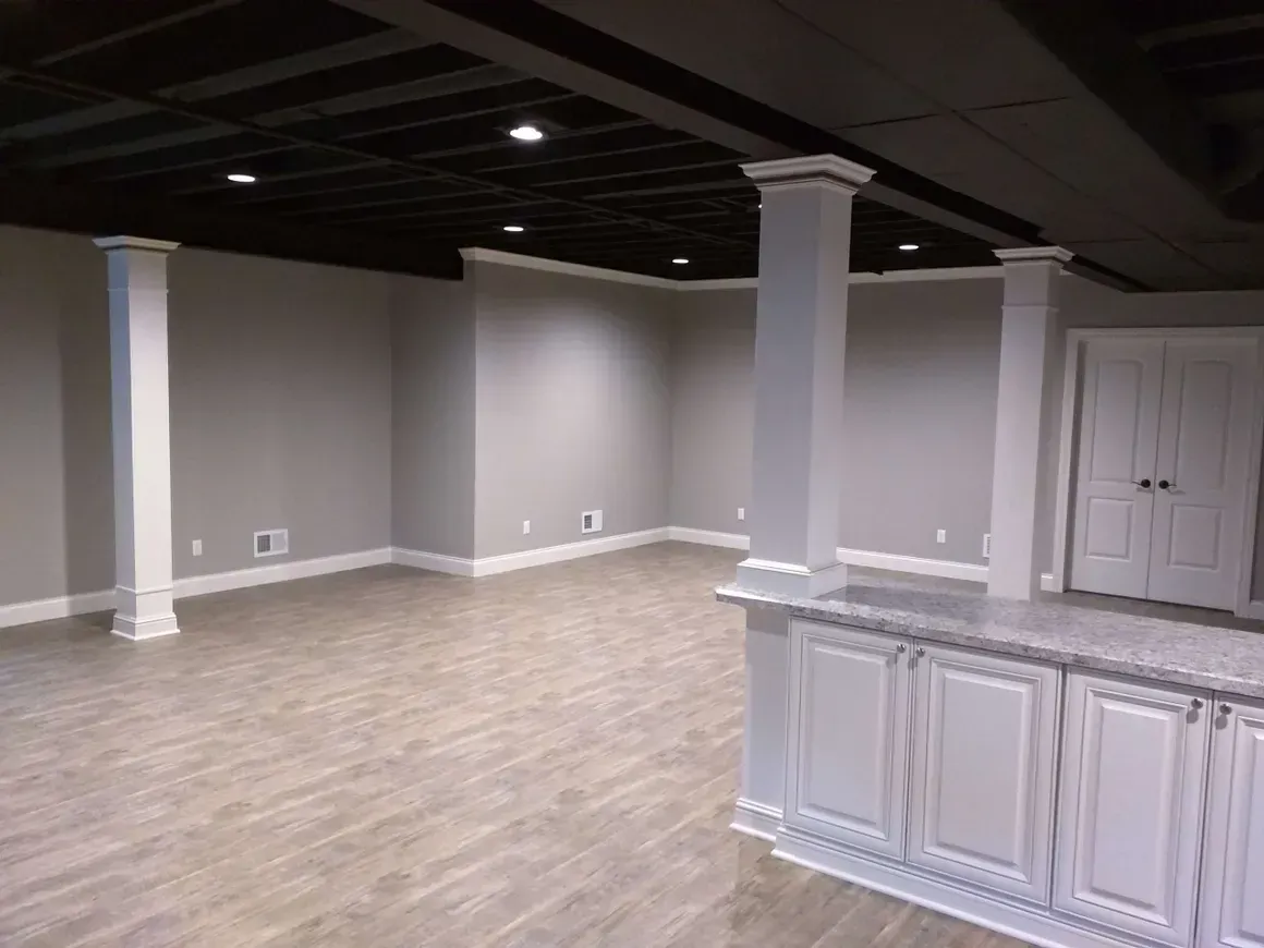 A finished basement with light gray walls, white trim, wood-look flooring, a black exposed ceiling, and white pillars.