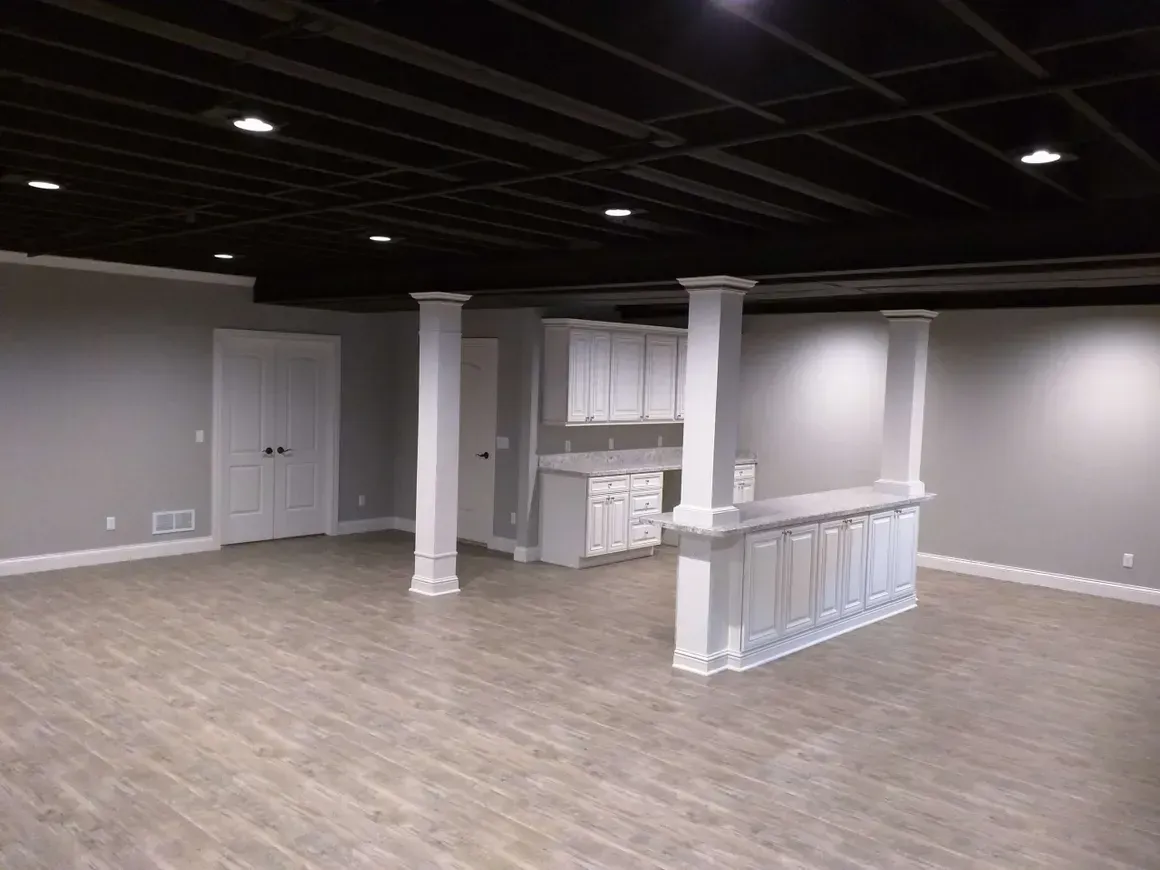 A basement with light gray walls, wood-look flooring, a black open-joist ceiling, and a white wet bar area with columns.