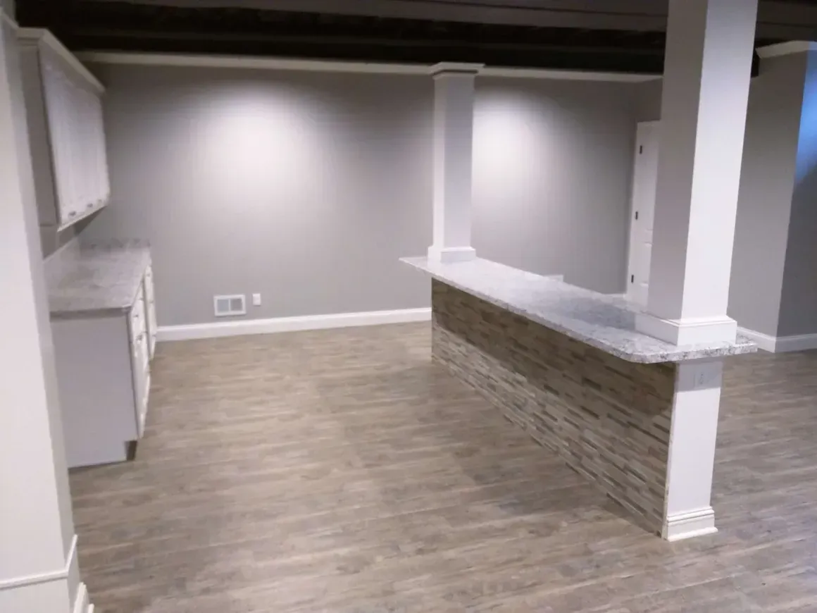 A modern basement kitchen featuring white cabinetry, a stone-front bar, and light wood-look tile flooring.