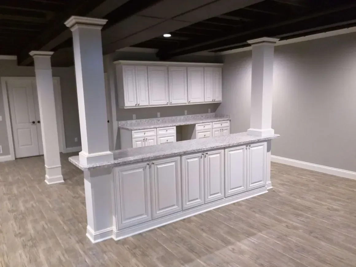A basement kitchenette with white cabinets, a matching bar counter, and support pillars on a light wood floor.