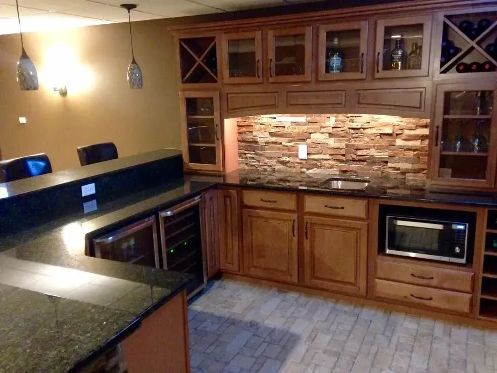 A home bar with wooden cabinets, dark countertops, a stone backsplash, a microwave, wine storage, and two pendant lights.