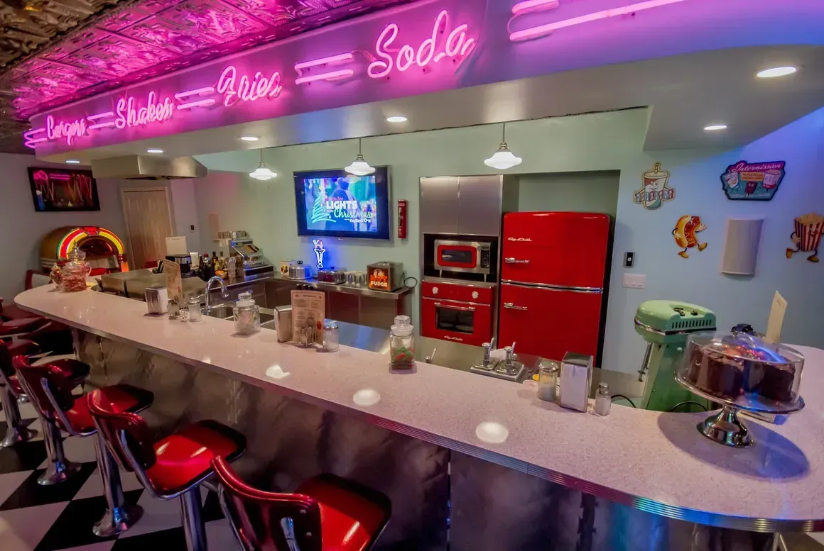 A retro-style home bar with a checkered floor, red vinyl stools, a pink neon sign, and a vintage-style red refrigerator.