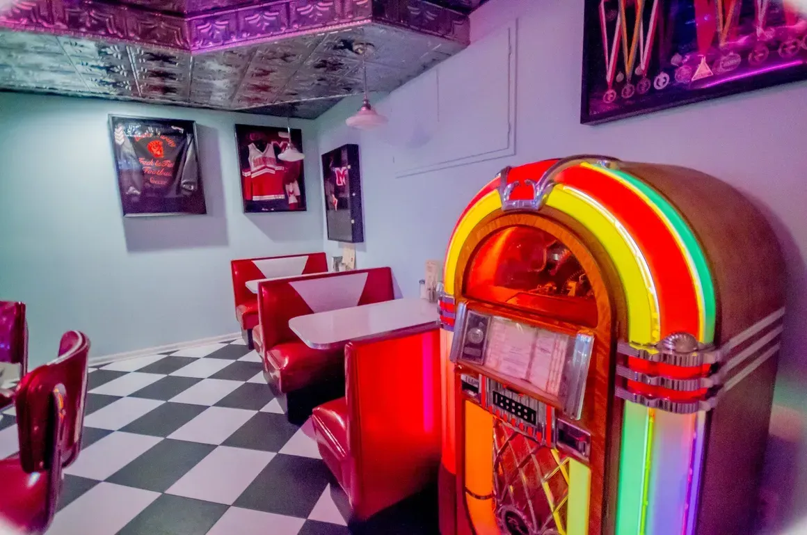 A 1950s-style diner booth area with a jukebox, checkered floor, red vinyl booths, and vintage wall posters.