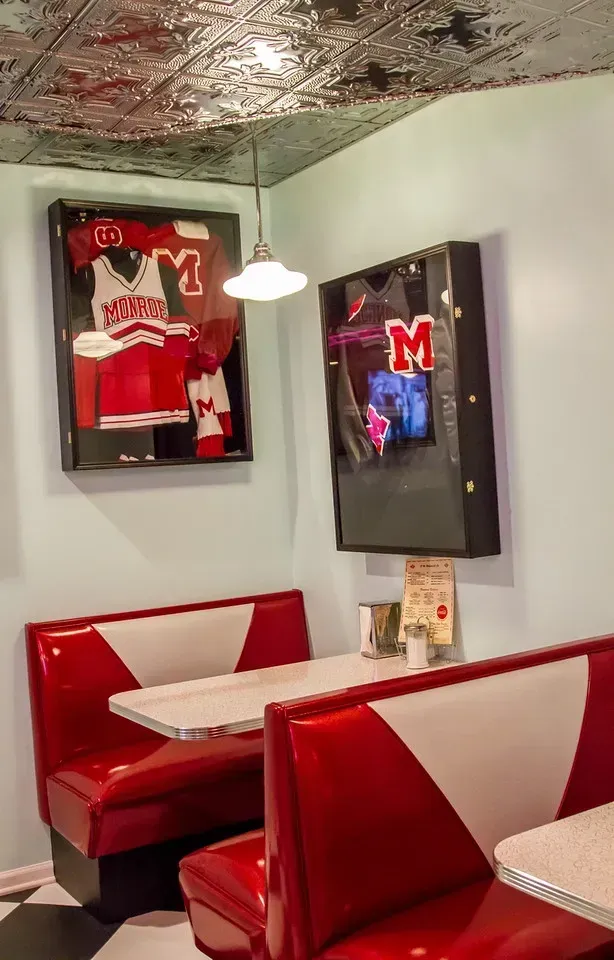A retro-style diner booth with red and white vinyl seats, checkerboard floors, and framed sports memorabilia on the wall.
