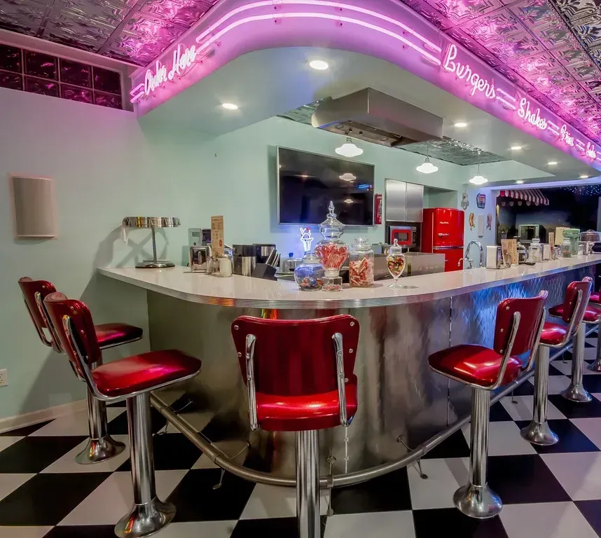 A retro-style home bar with a checkered floor, red vinyl stools, light blue walls, and a neon-lit counter.