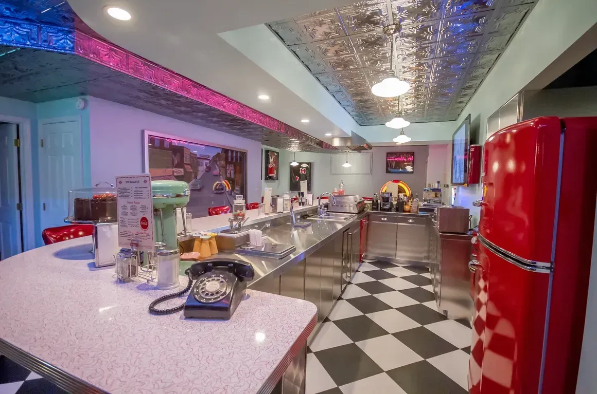 Retro 1950s-style kitchen with a checkerboard floor, red refrigerator, pink granite counters, and metallic ceiling.