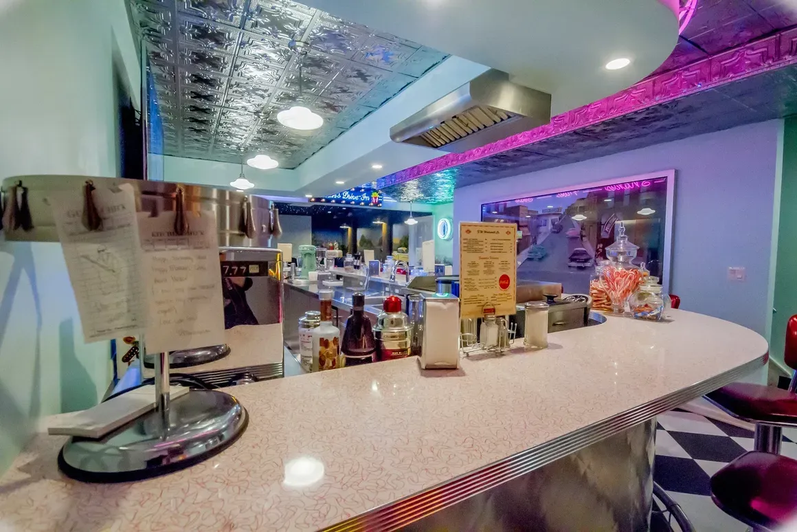 A brightly lit, retro-style diner counter with a speckled pink countertop, checkered flooring, and silver ceiling tiles.