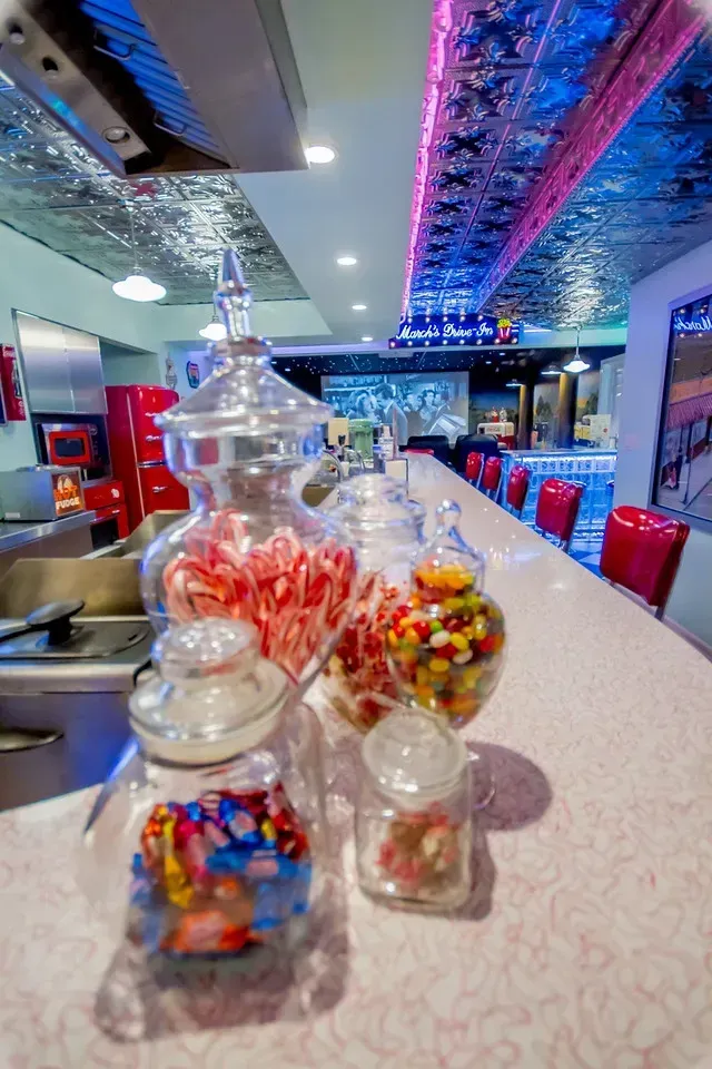 Four glass jars filled with various candies sit on a pink diner counter, with red booths visible in the background.