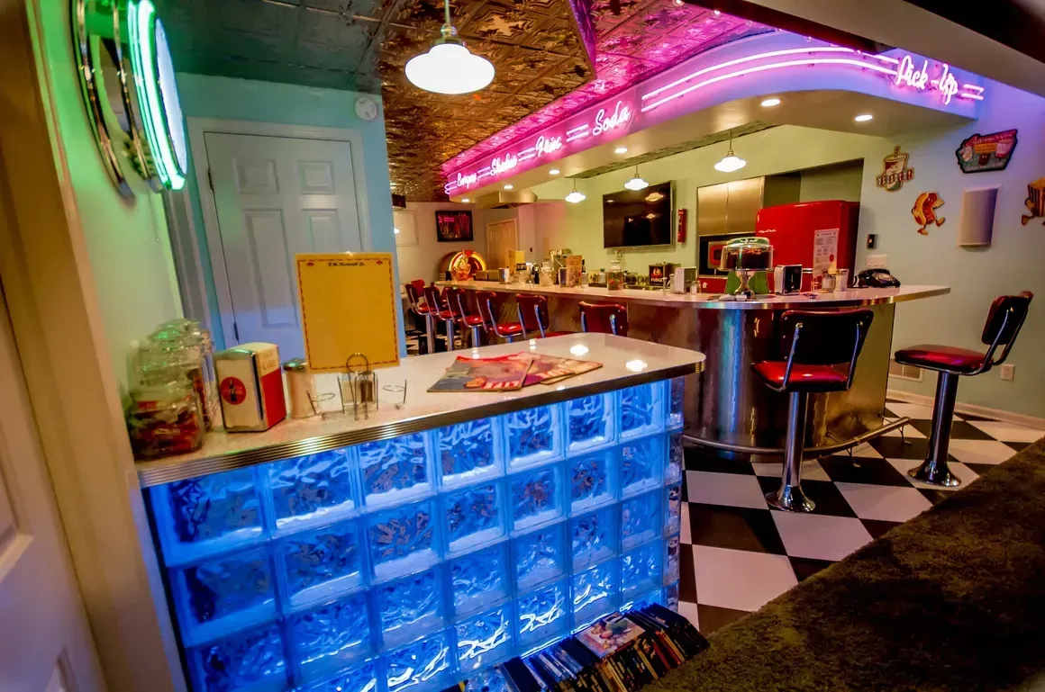 A retro-style basement diner with a glowing blue glass-block bar, neon lights, and checkerboard flooring.