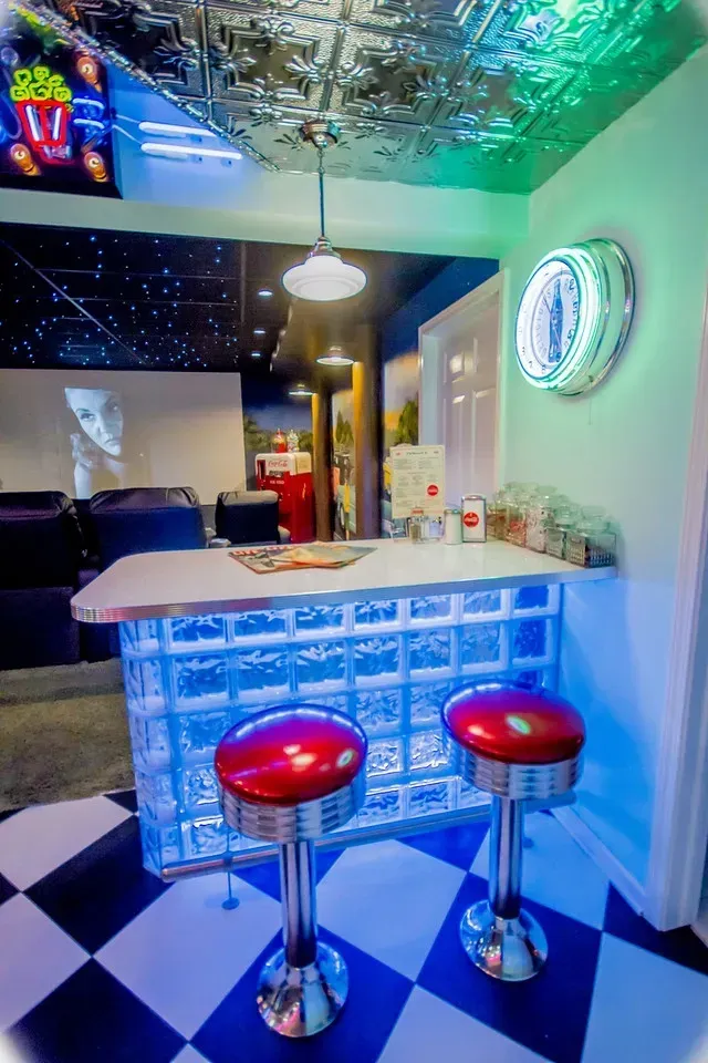 A retro-style home bar with two red stools, a glass-block counter, and blue lighting, set in a room with a checkerboard floor.