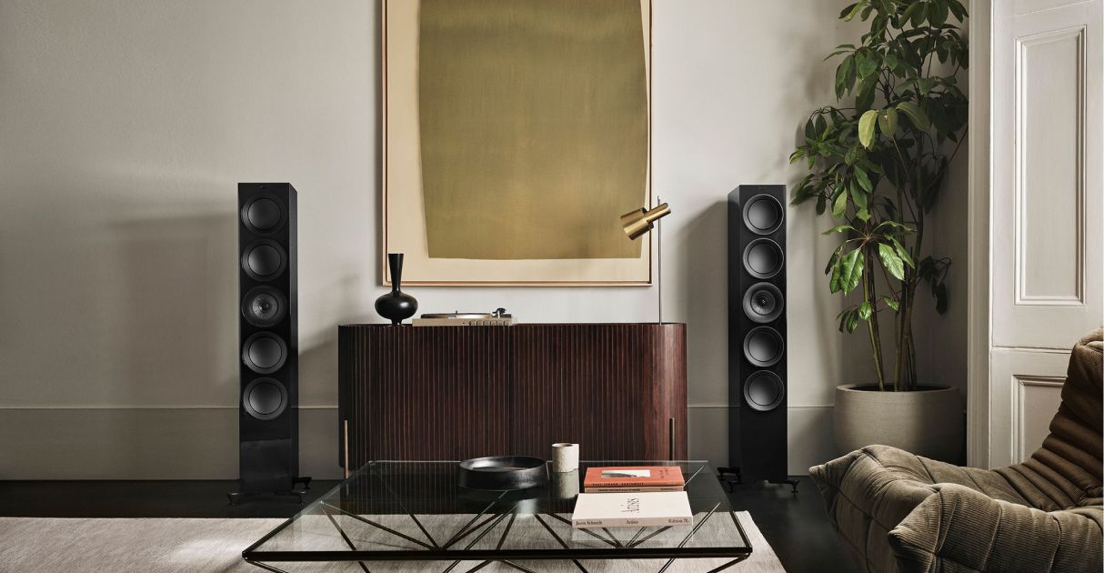 A living room with a coffee table , chairs , speakers and a plant.