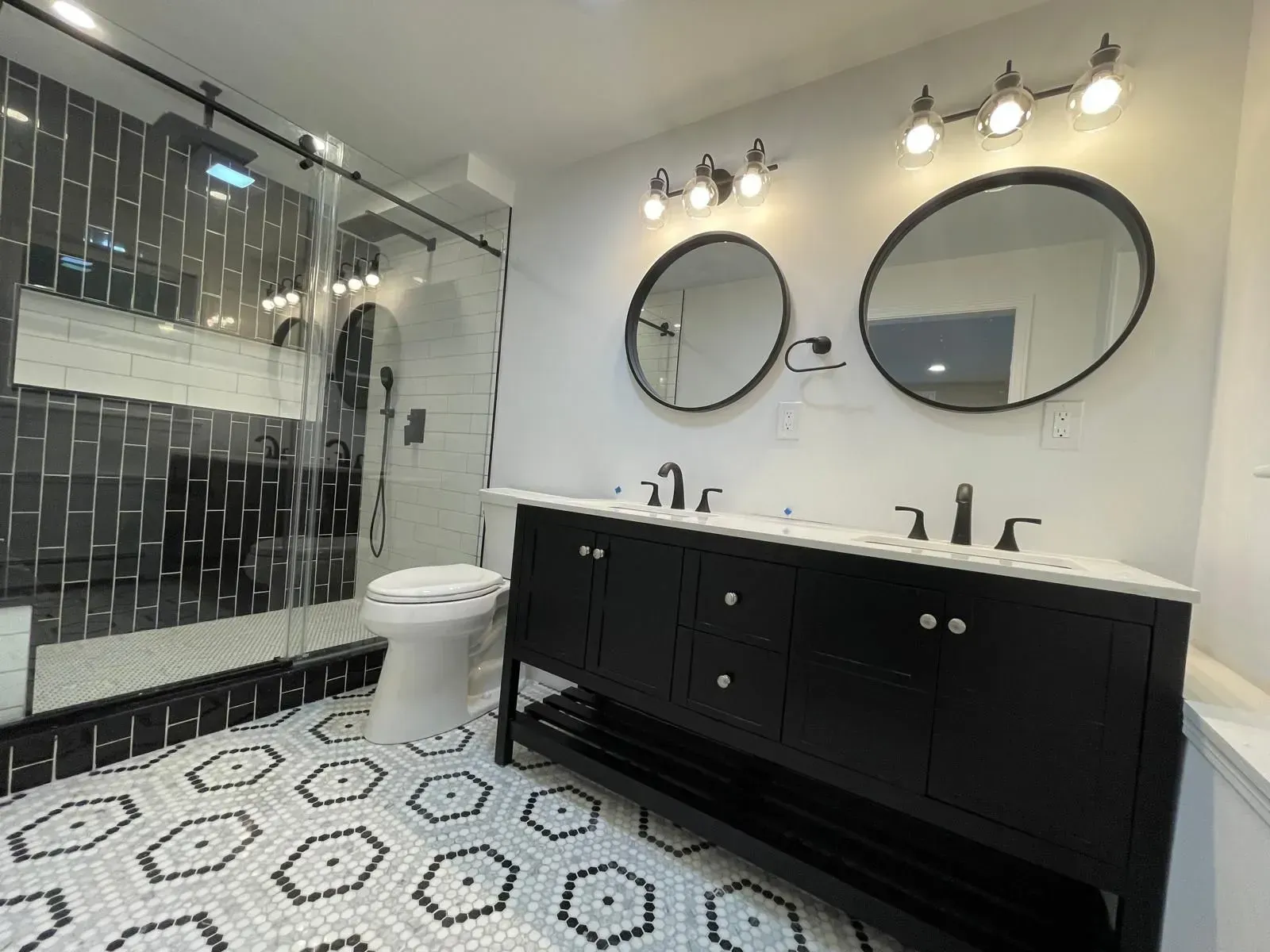 Black and white bathroom with hexagonal patterned floor, dark vanity, and two round mirrors.