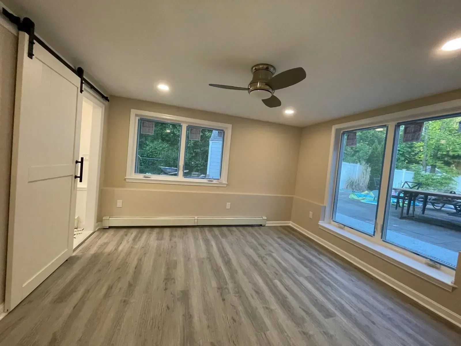 Room with light gray wood flooring, beige walls, windows, and a sliding white door.