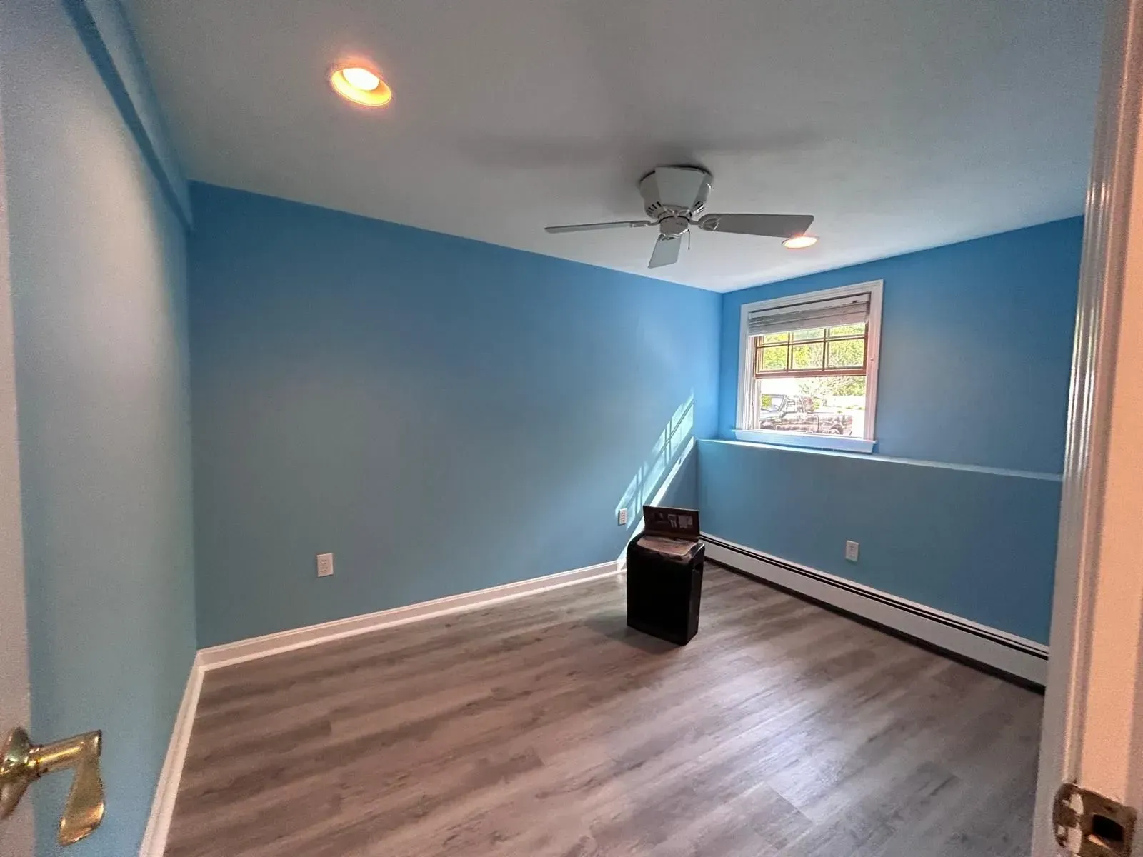 Empty room with blue walls, gray flooring, window, and ceiling fan. Black object sits in center.