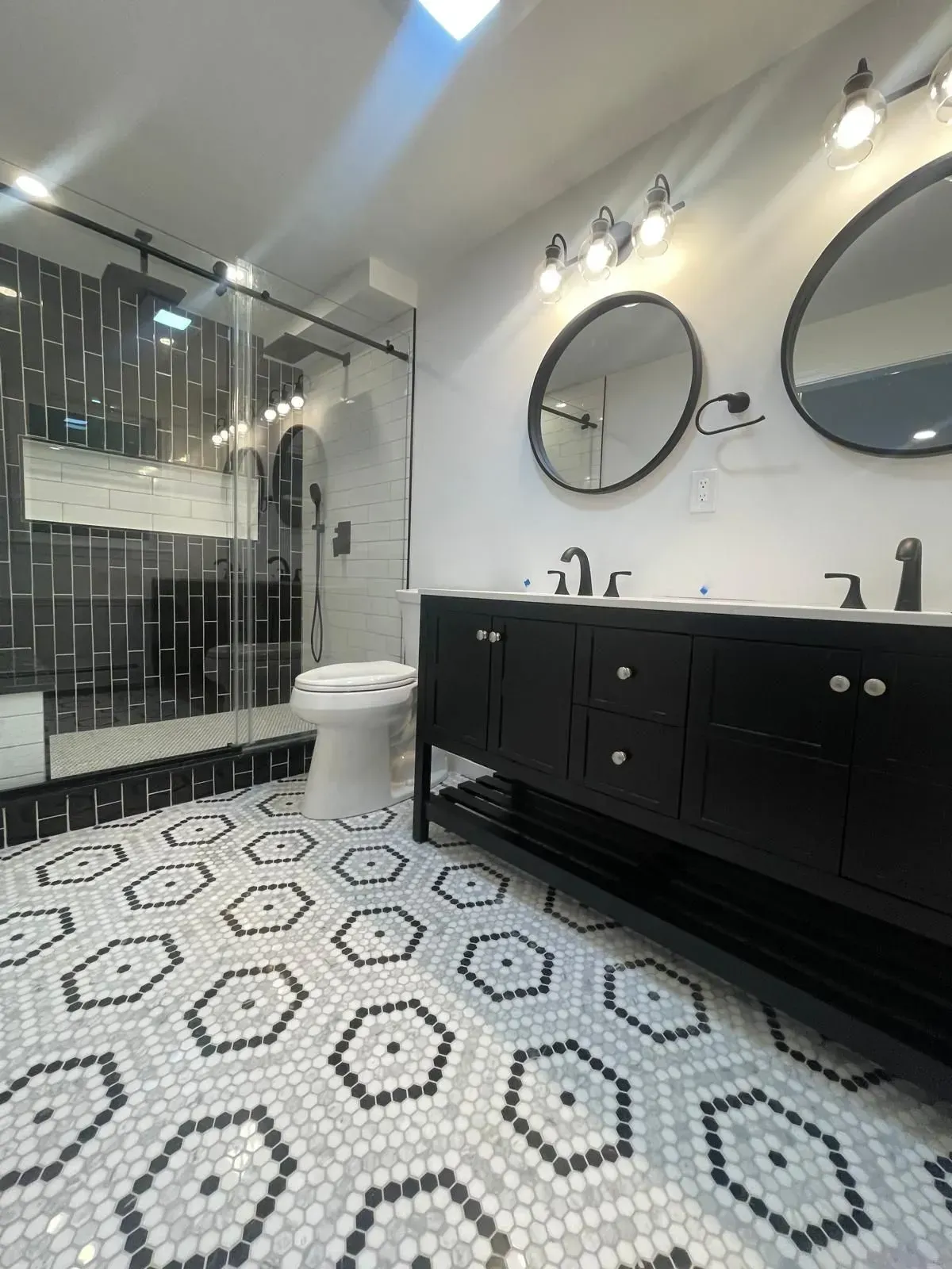 Modern bathroom with black vanity, round mirrors, geometric tile floor, and glass shower.