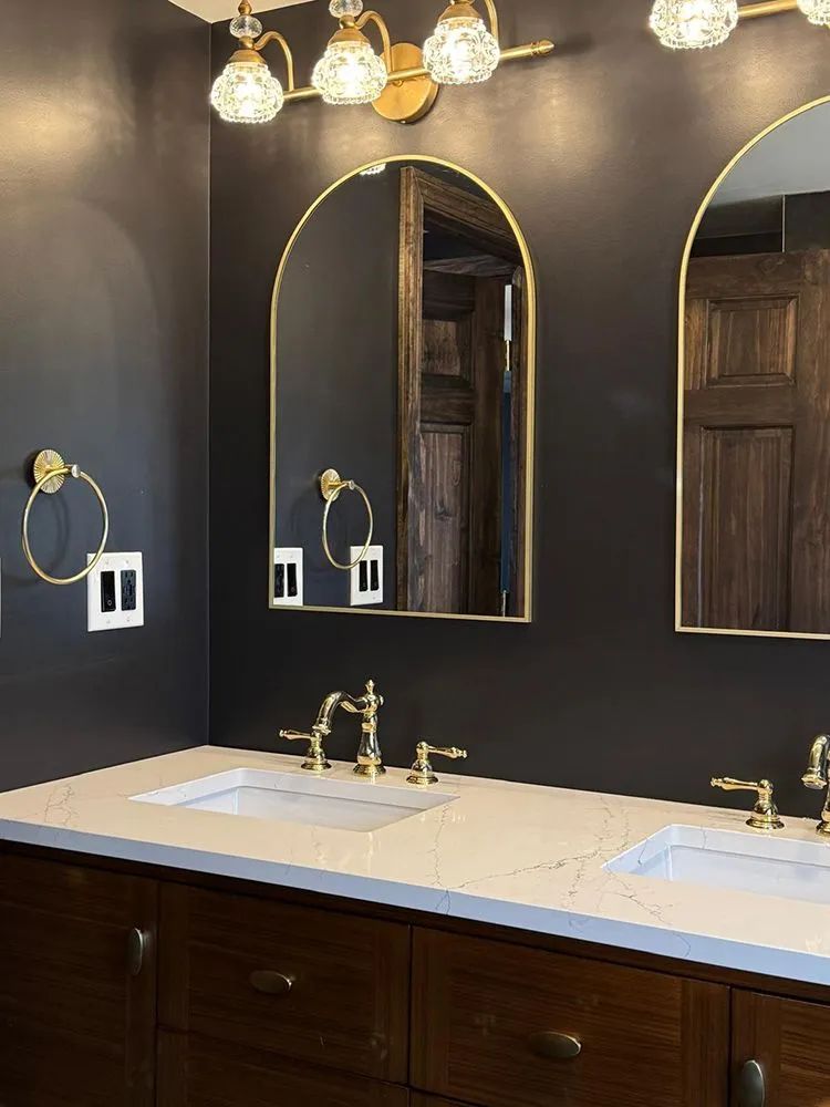Bathroom with gold fixtures, dark gray walls, arched mirrors, and wood cabinetry.