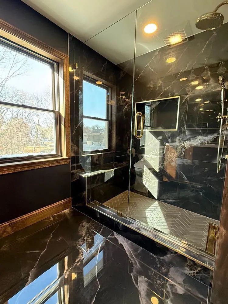 Luxurious bathroom with black marble walls and floor, gold fixtures, large windows, and a glass shower.