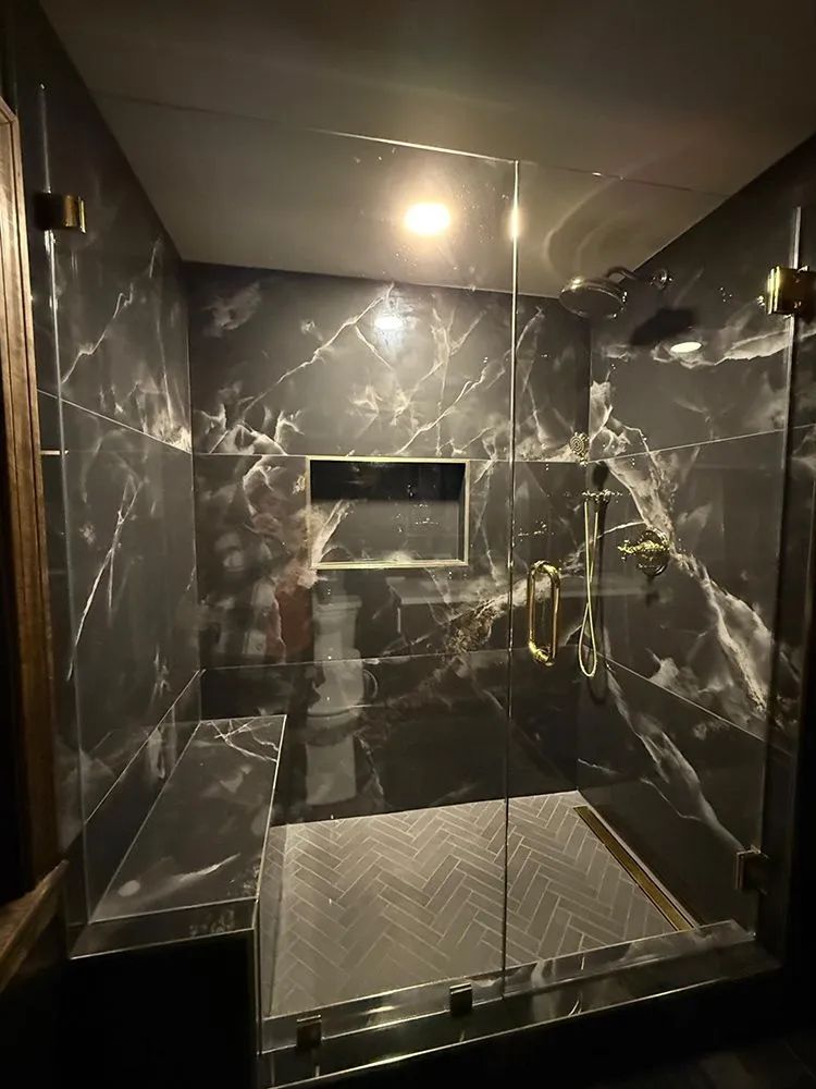 Luxurious shower with black marble walls, gold fixtures, and a glass door. Bench and shelf included.