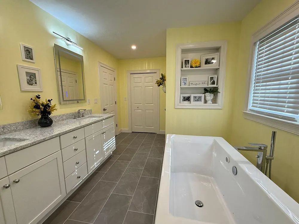 Bathroom with yellow walls, white cabinets, and a gray tile floor. A bathtub sits near a window.