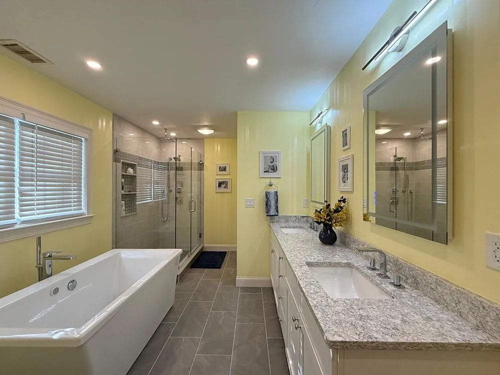 Bathroom with yellow walls, white tub, gray floor, granite countertop, and glass shower.
