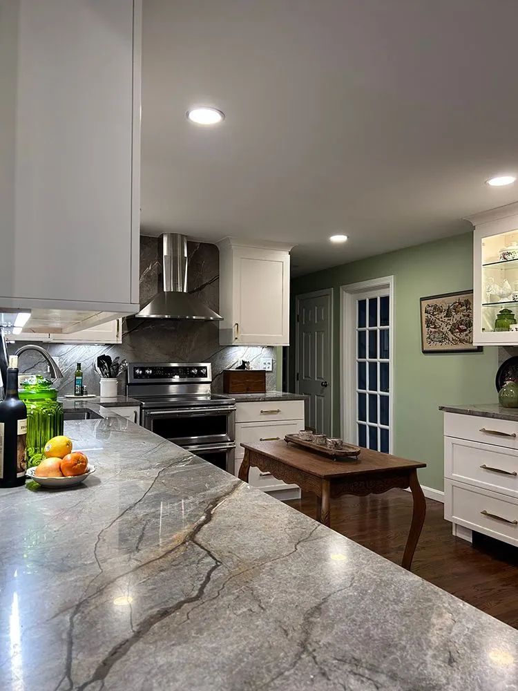 Kitchen with light cabinets, gray countertops, and stainless steel appliances. A small wooden table and sage green wall are visible.