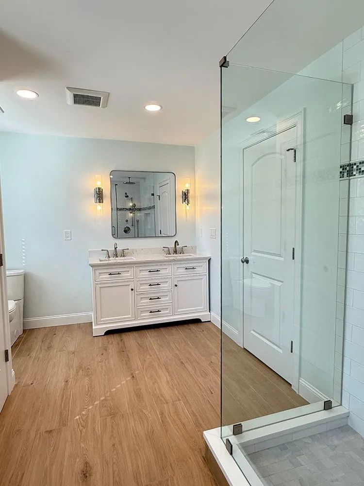 Bathroom with a white double vanity, light blue walls, wood-look floor, and a glass shower.