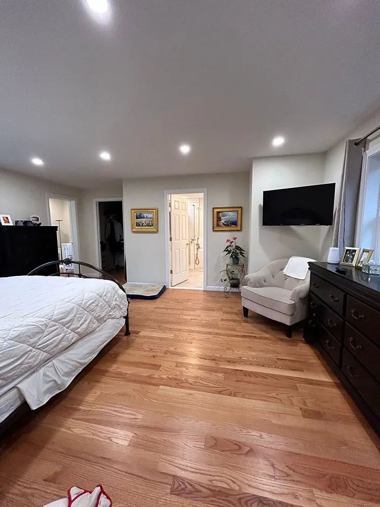Bedroom with hardwood floors, bed, dresser, chair, TV, and open doorway to bathroom.