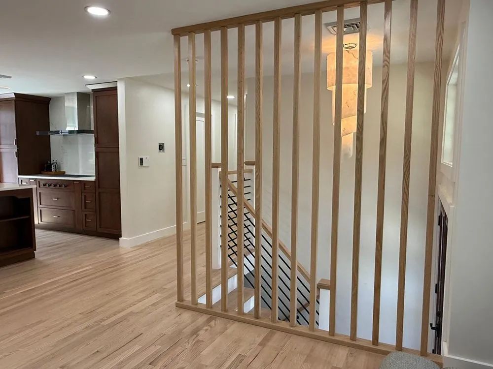 Wooden slat room divider separates a staircase from an entryway with hardwood floors and kitchen in the background.