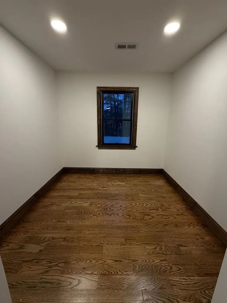 Empty room with dark hardwood floor, white walls, small window, and recessed lighting.