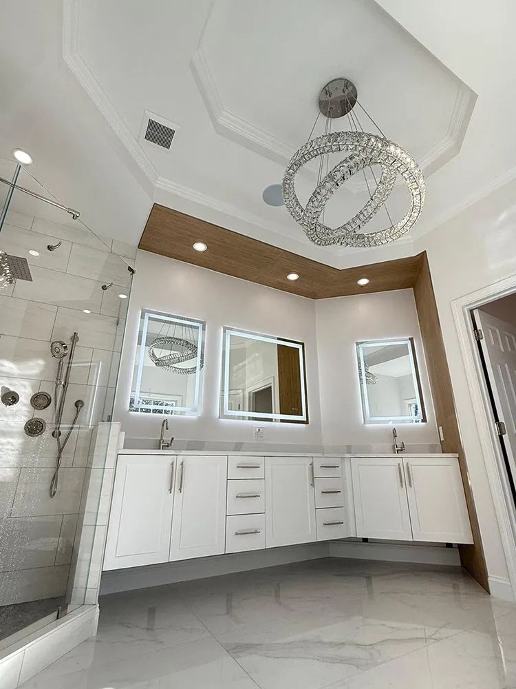 Modern white bathroom with double vanity, illuminated mirrors, and a sparkling chandelier.