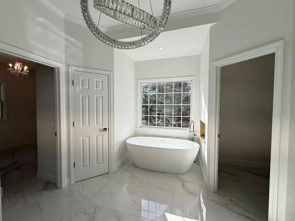 Luxurious white bathroom with a freestanding tub, window, and large chandelier.