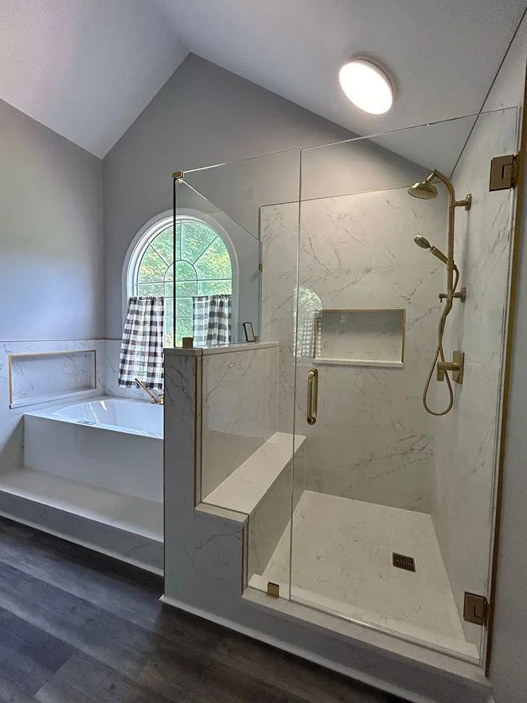 Bathroom with glass shower, white marble tile, gold fixtures, and a soaking tub with a window.