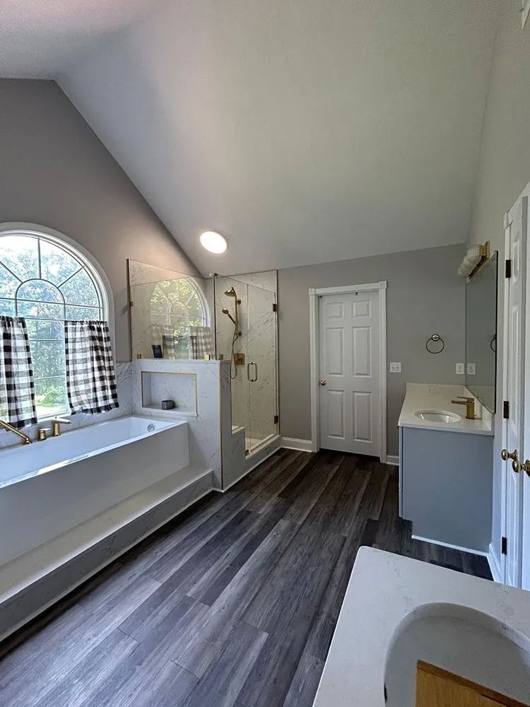 Spacious bathroom with gray walls, wood-look flooring, large shower, bathtub by window with plaid curtains.