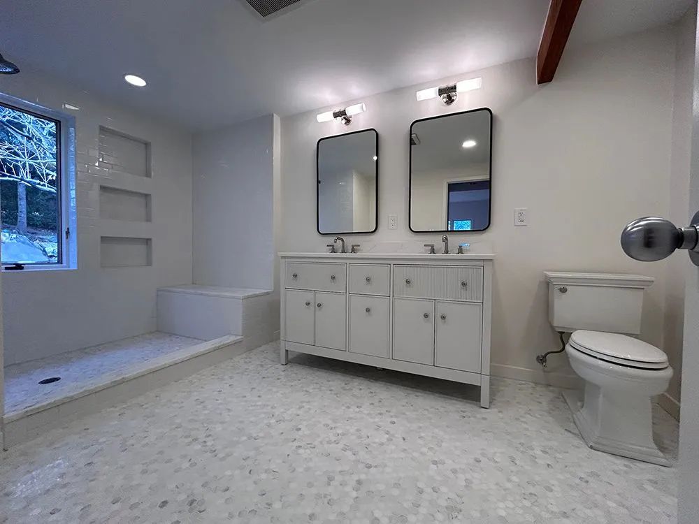 White bathroom with vanity, mirrors, and a toilet. Pebble tile floor. Window and built-in shower.