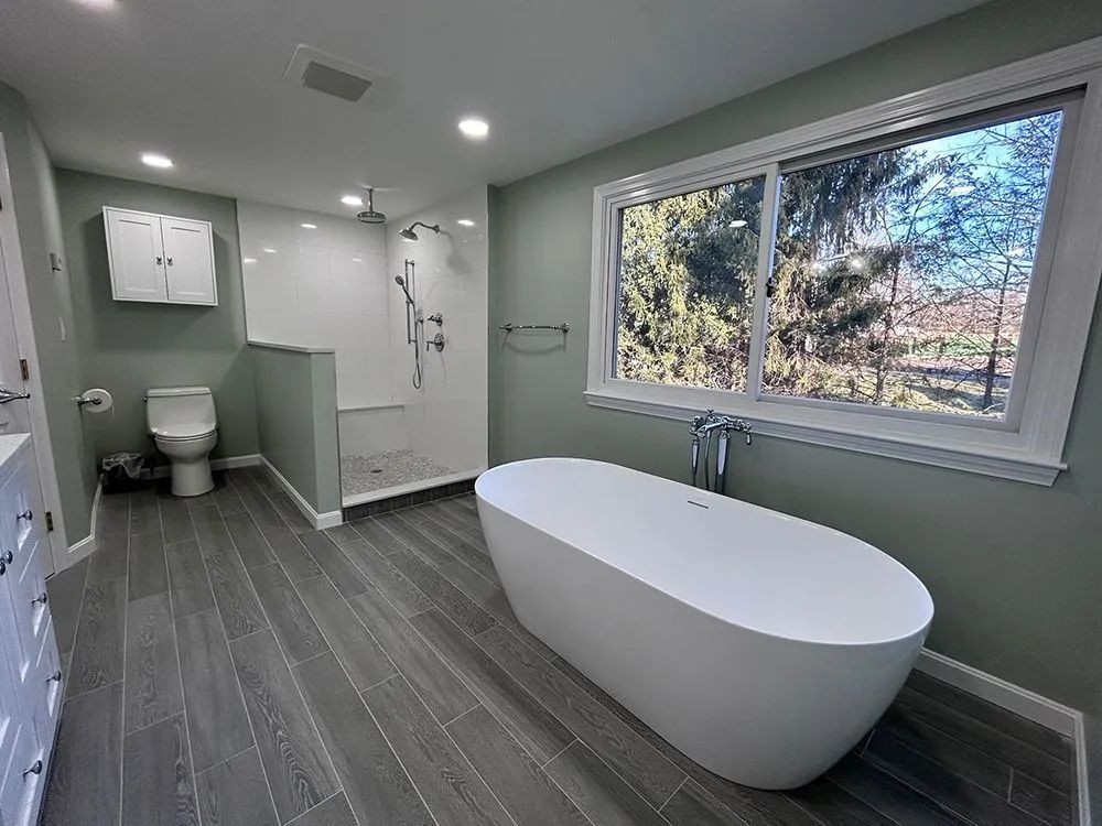 Modern bathroom with a white tub, shower, and toilet, gray wood-look tile, and a large window.