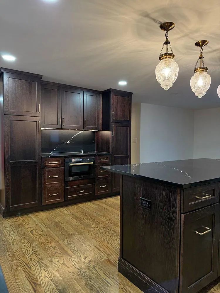 Dark wood kitchen with cabinetry, island, and pendant lights. Hardwood floors and black countertop.