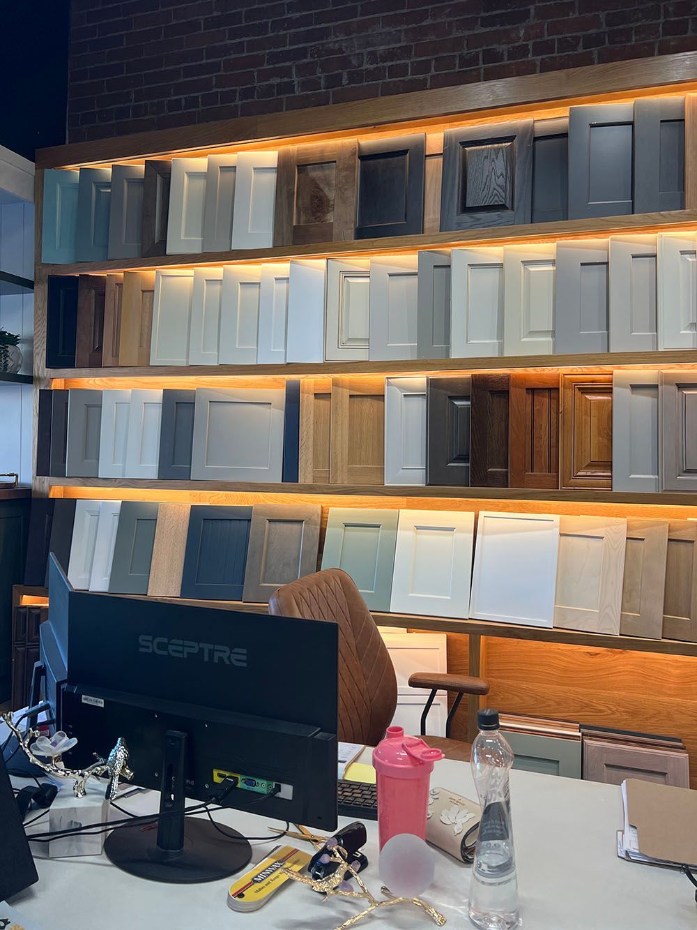 Office desk with monitor and cups in front of a wall of gray and white shutters lit by warm shelves
