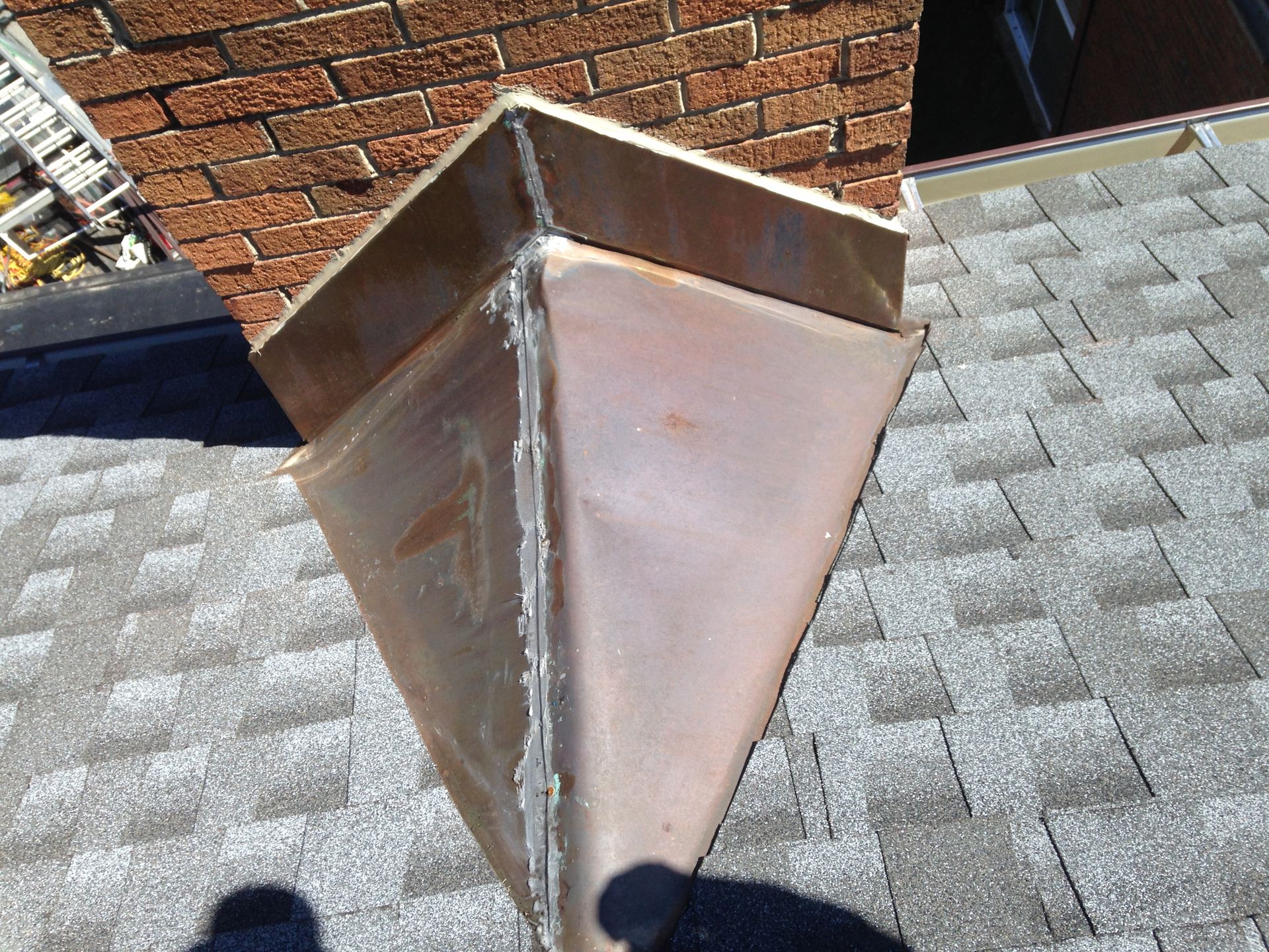 Metal roof vent on a shingled rooftop beside a brick wall, with visible seams and rust stains