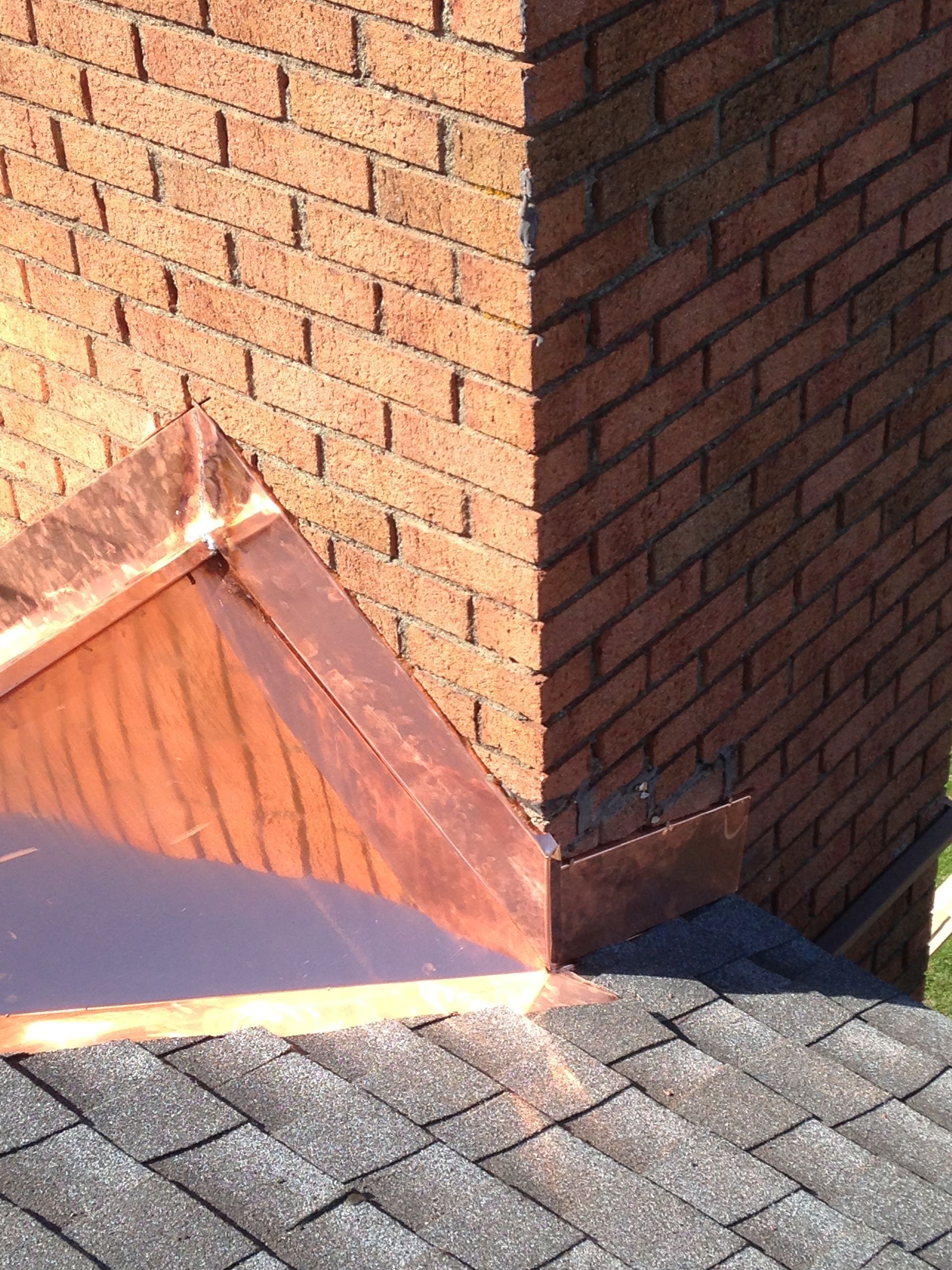 Brick chimney on a shingled roof with a copper flashing base in sunlight