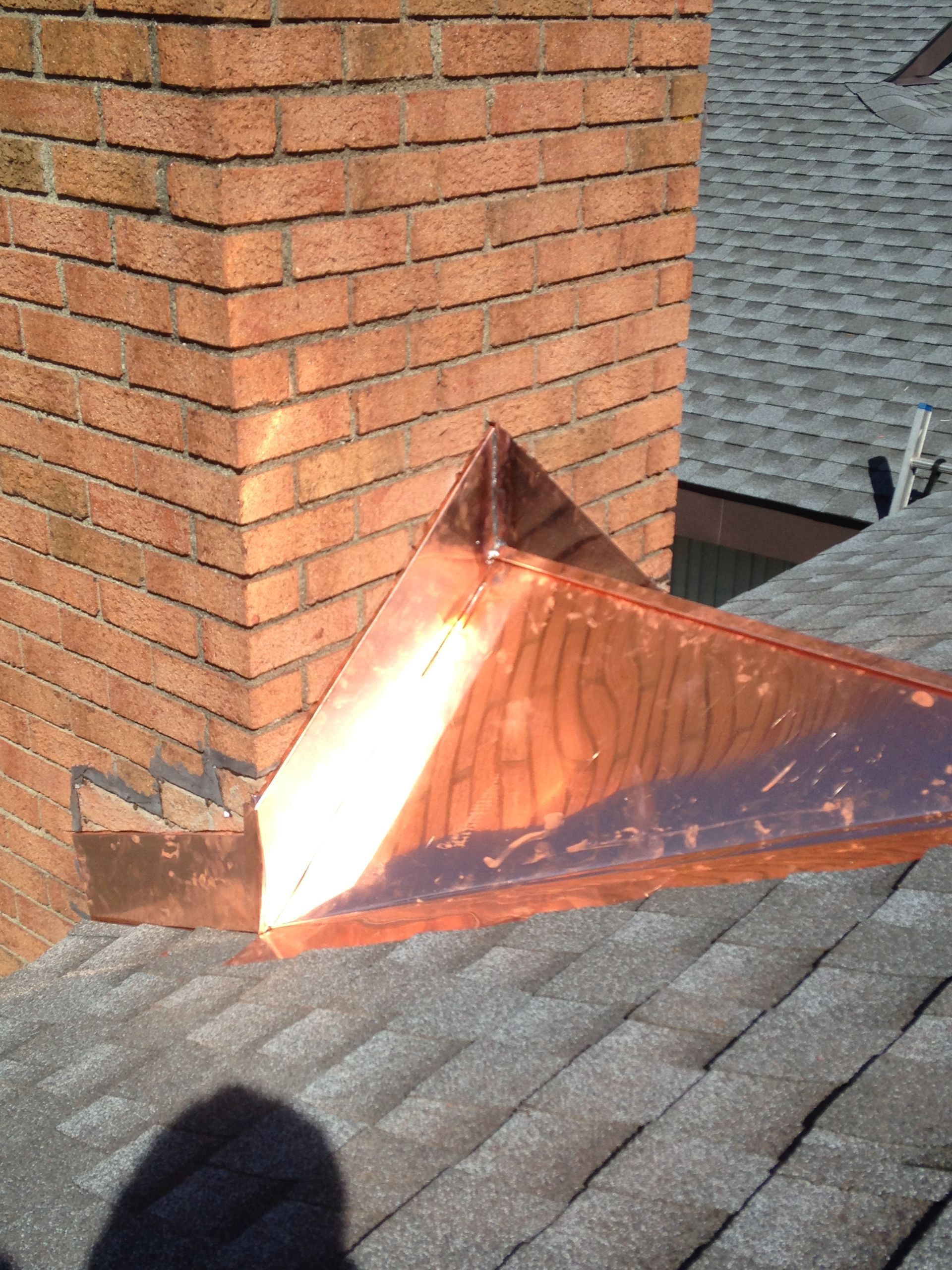 Copper chimney flashing on a shingled roof beside a brick chimney.